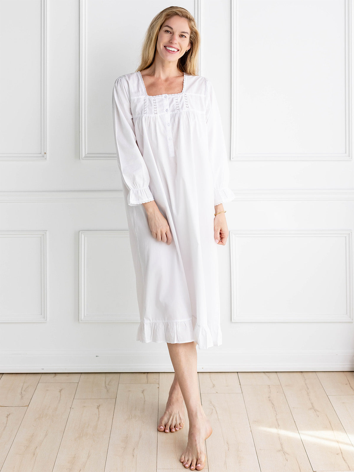 Woman wearing a white nightgown standing in a room with white walls and wooden floor.