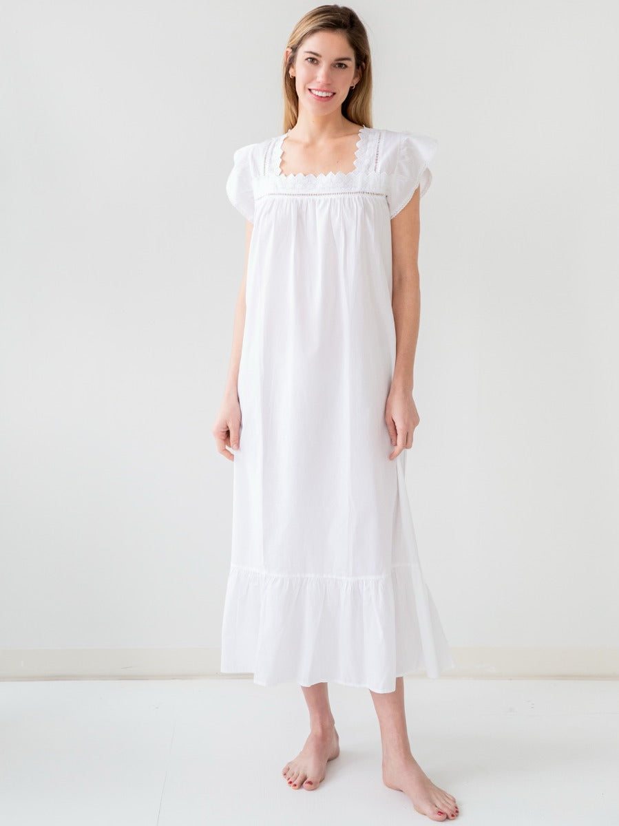 A woman standing in a white Genevieve white cotton nightgown with butterfly cap sleeves and a scalloped square neckline.