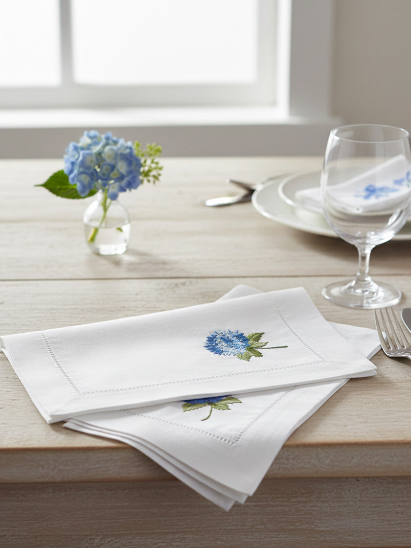 White napkins with floral embroidery on a wooden table with a vase of blue flowers and glasses.