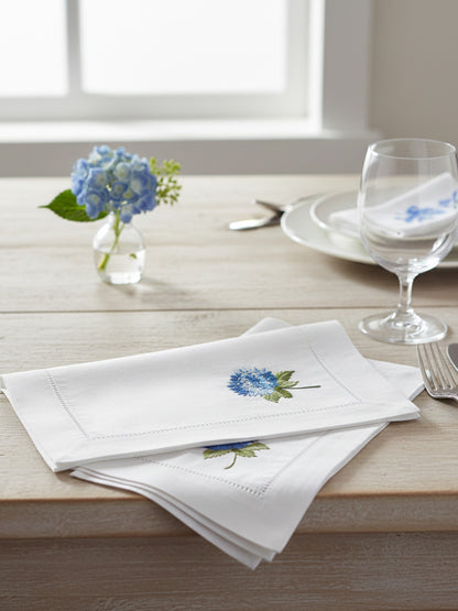 White napkins with floral embroidery on a wooden table with a vase of blue flowers and glasses.