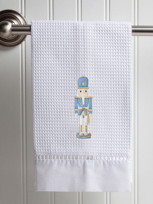 A white waffle-weave kitchen towel hanging from a metal rod on a white paneled door. The towel features a central embroidered nutcracker figure in blue, gold, and white, with a solid white ruffled hem.