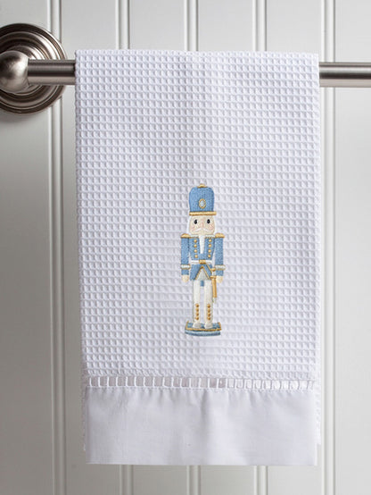 A white waffle-weave kitchen towel hanging from a metal rod on a white paneled door. The towel features a central embroidered nutcracker figure in blue, gold, and white, with a solid white ruffled hem.