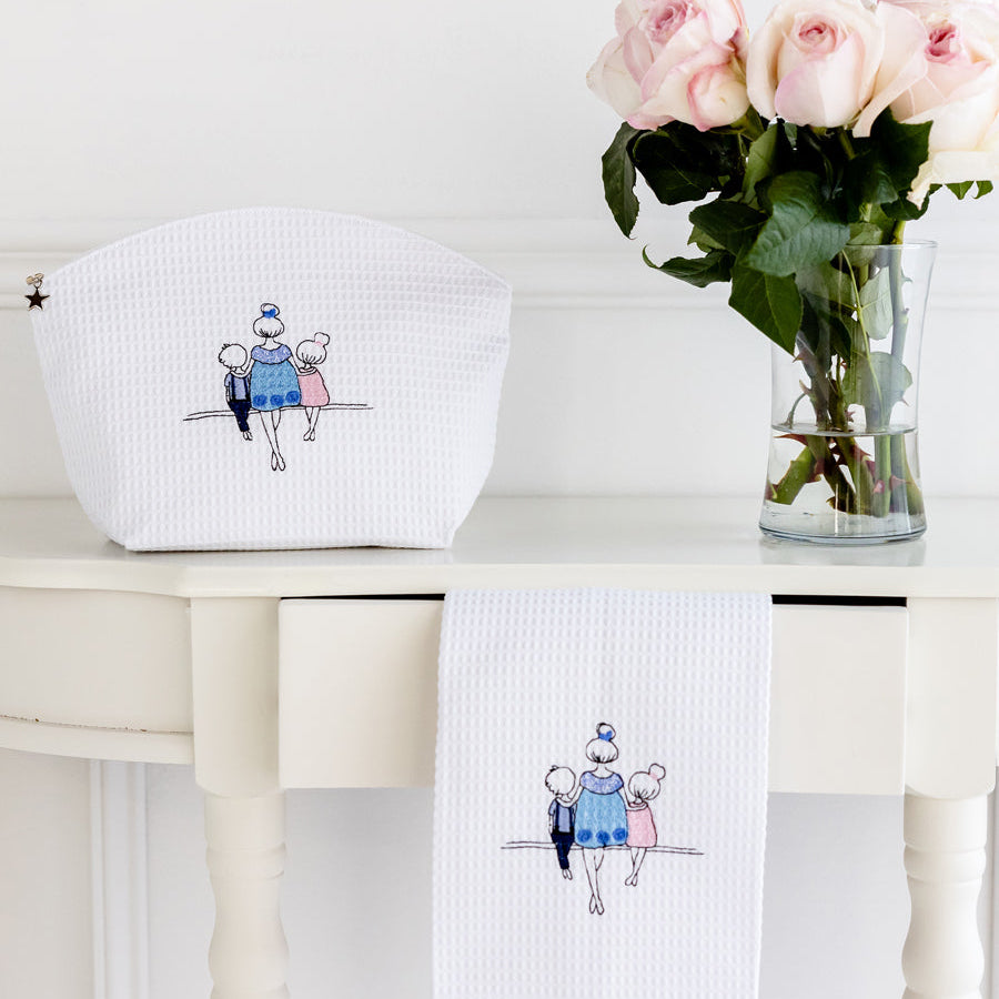 White pouch and towel with embroidered design on a white surface, accompanied by a vase of pink roses.
