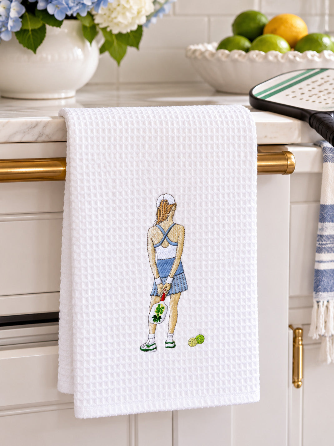 A white waffle-weave kitchen towel with pickle ball girl design hanging from a gold towel bar on a white kitchen counter. The towel features an embroidered illustration of a pickle ball player from behind holding a racket, with tennis balls near her feet.