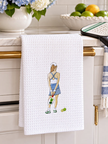 A white waffle-weave kitchen towel with pickle ball girl design hanging from a gold towel bar on a white kitchen counter. The towel features an embroidered illustration of a pickle ball player from behind holding a racket, with tennis balls near her feet.
