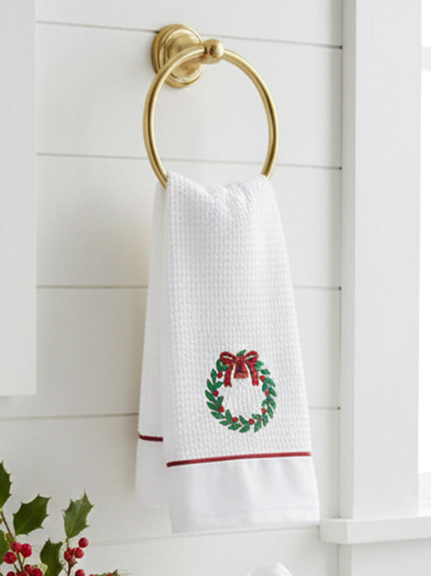 White towel with a Christmas wreath design hanging on a gold towel ring against a white wooden wall.