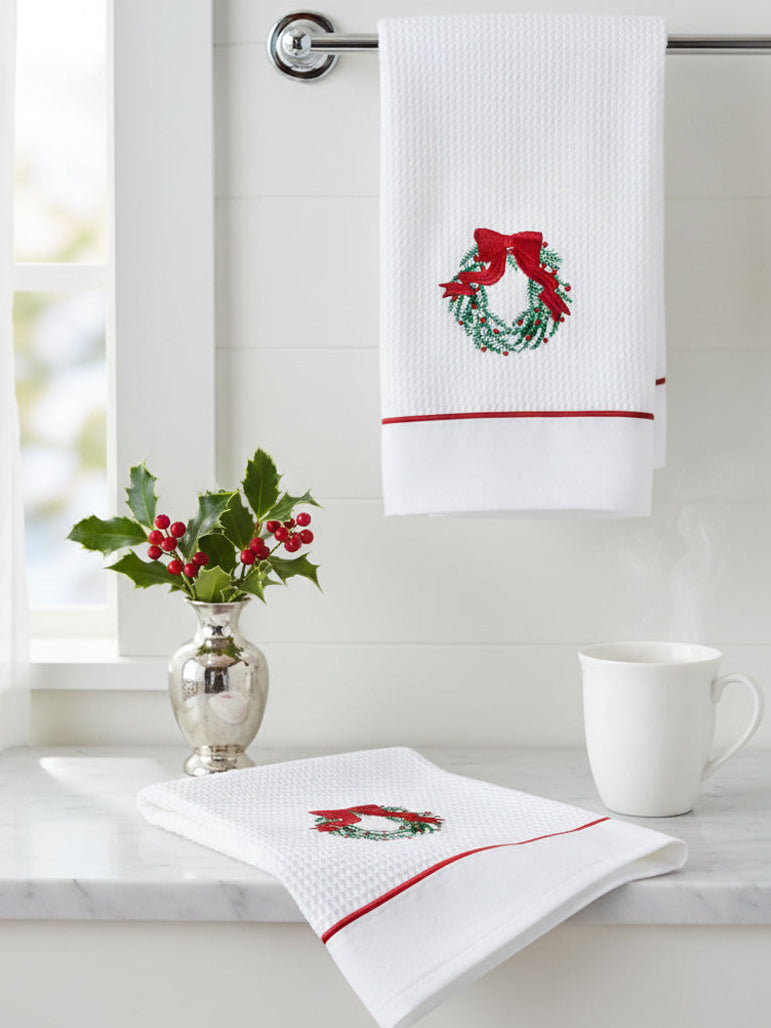 White towel with embroidered wreath design hanging on a rack, with a cup and decorative plant on a surface below.