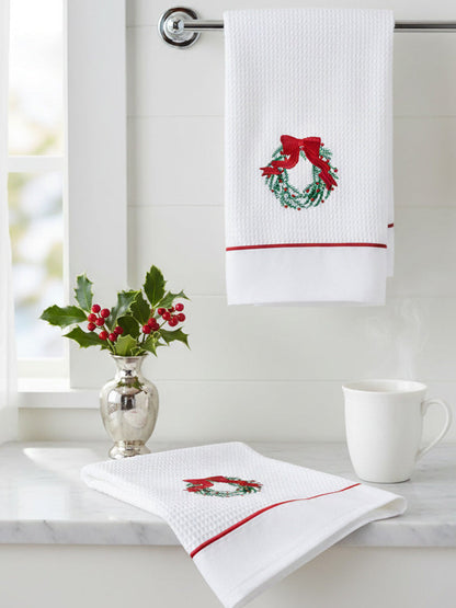 White towel with embroidered wreath design hanging on a rack, with a cup and decorative plant on a surface below.