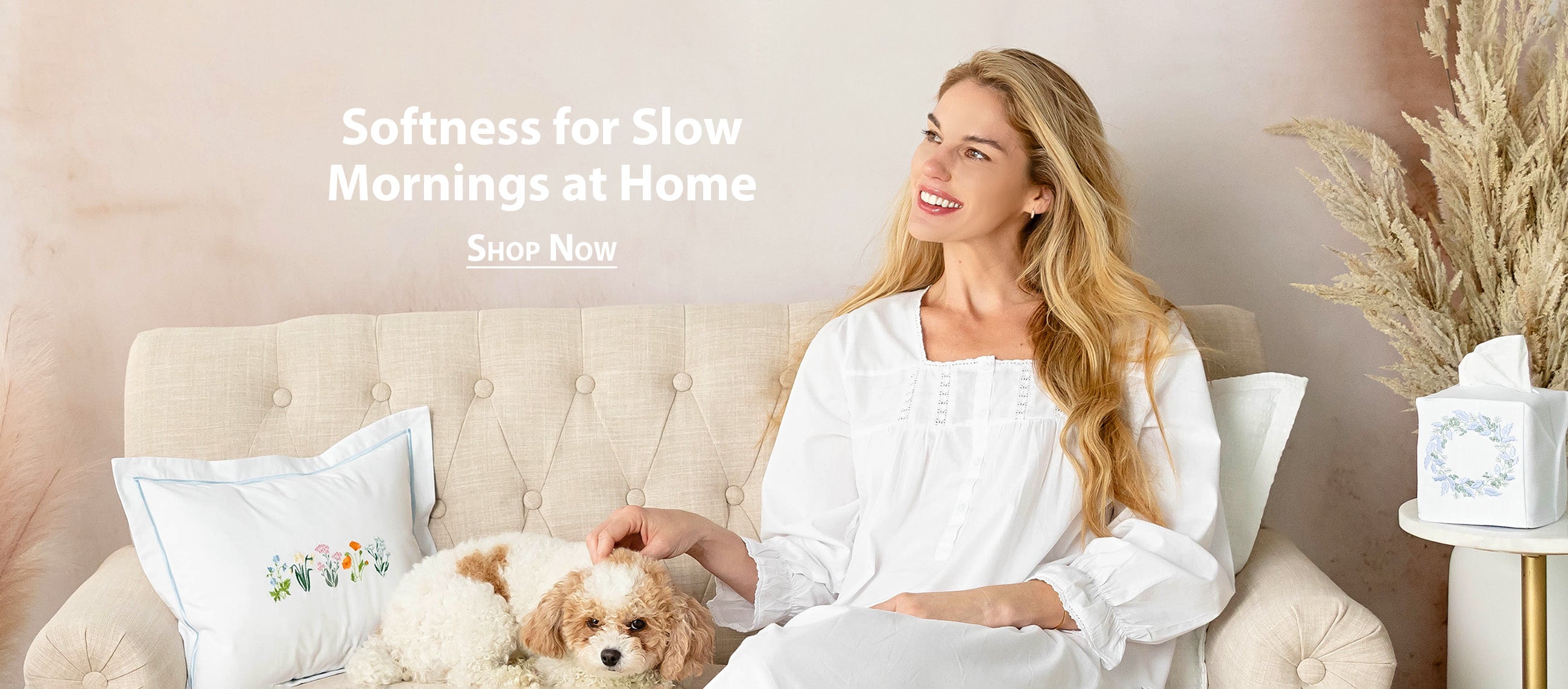Woman in a white nightgown from Jacaranda Living sitting on a couch with a dog, surrounded by soft pillows.