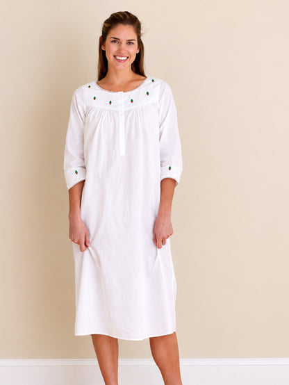 A woman modeling a white long-sleeve cotton nightgown with small green embroidered details on the neckline and cuffs, set against a plain beige wall.