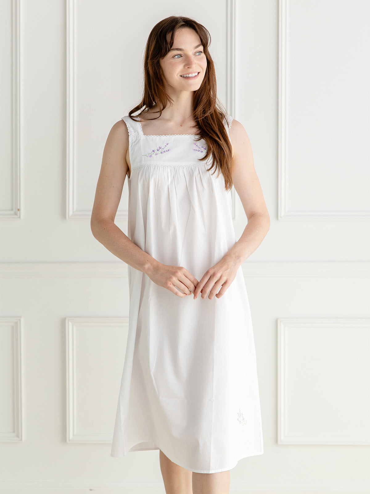 A woman wearing a white sleeveless nightgown with delicate embroidery on the chest, standing against a white paneled wall.