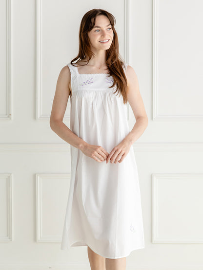 A woman wearing a white sleeveless nightgown with delicate embroidery on the chest, standing against a white paneled wall.