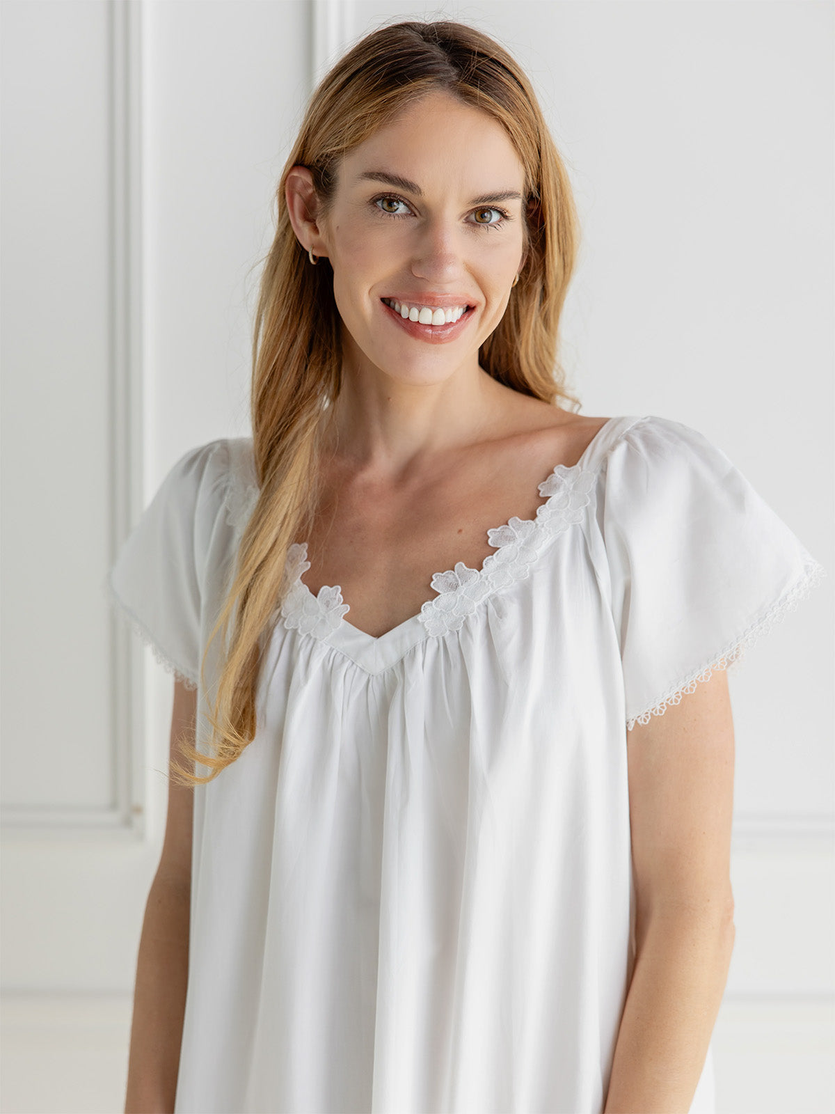 A smiling woman with long light brown hair wearing a white short-sleeve nightgown with a scalloped lace V-neckline, set against a bright, minimalist white background.