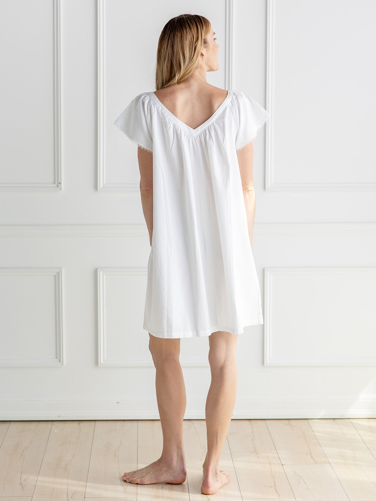 A woman stands barefoot in a bright white room, viewed from behind, wearing a loose white short-sleeve nightgown or tunic dress.