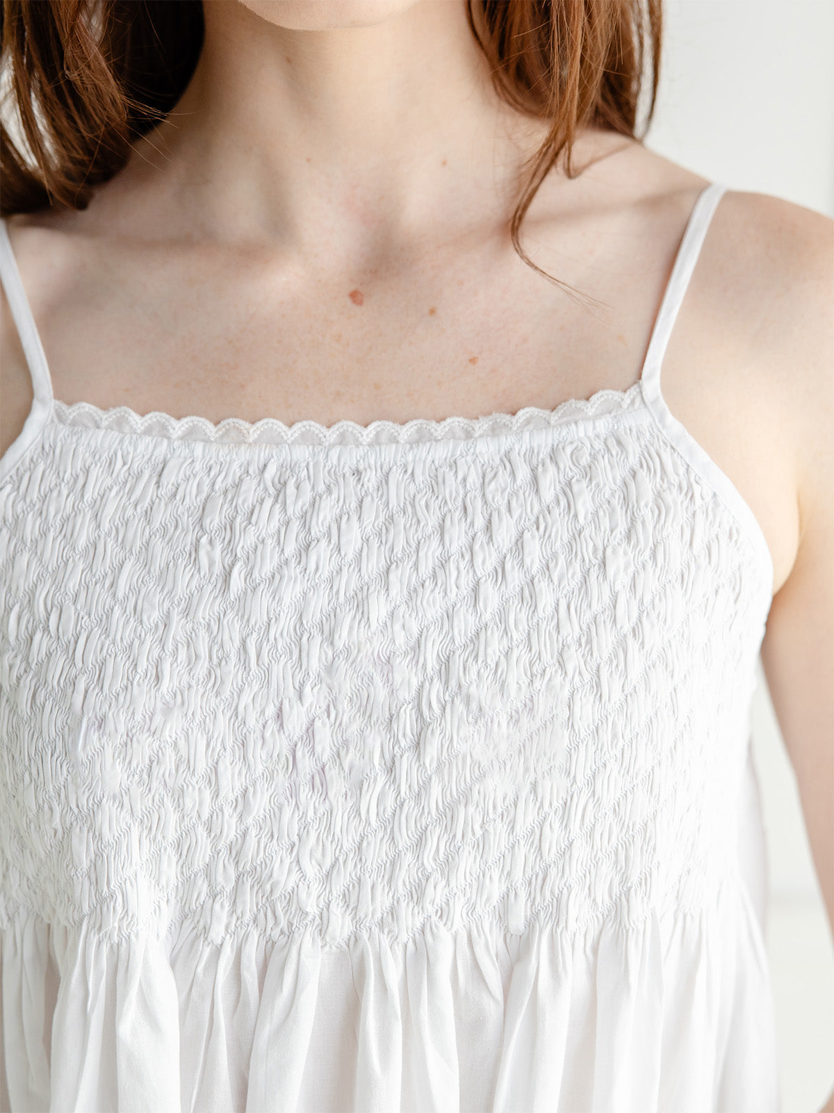 Close-up of a person wearing a white smocked camisole top with thin straps and a scalloped neckline.