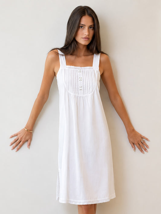 A woman modeling a white sleeveless midi dress with a square neckline, gathered bust, and decorative button details, posed against a plain light beige wall.