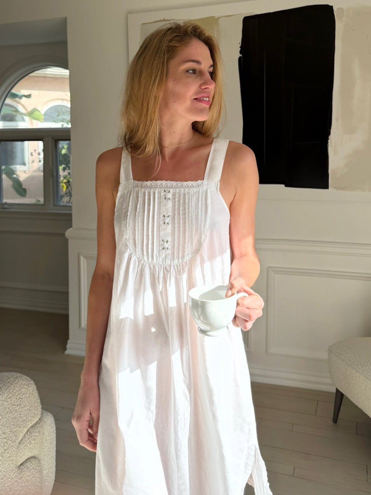 Woman in a white nightgown holding a cup in a room with light-colored walls and furniture.