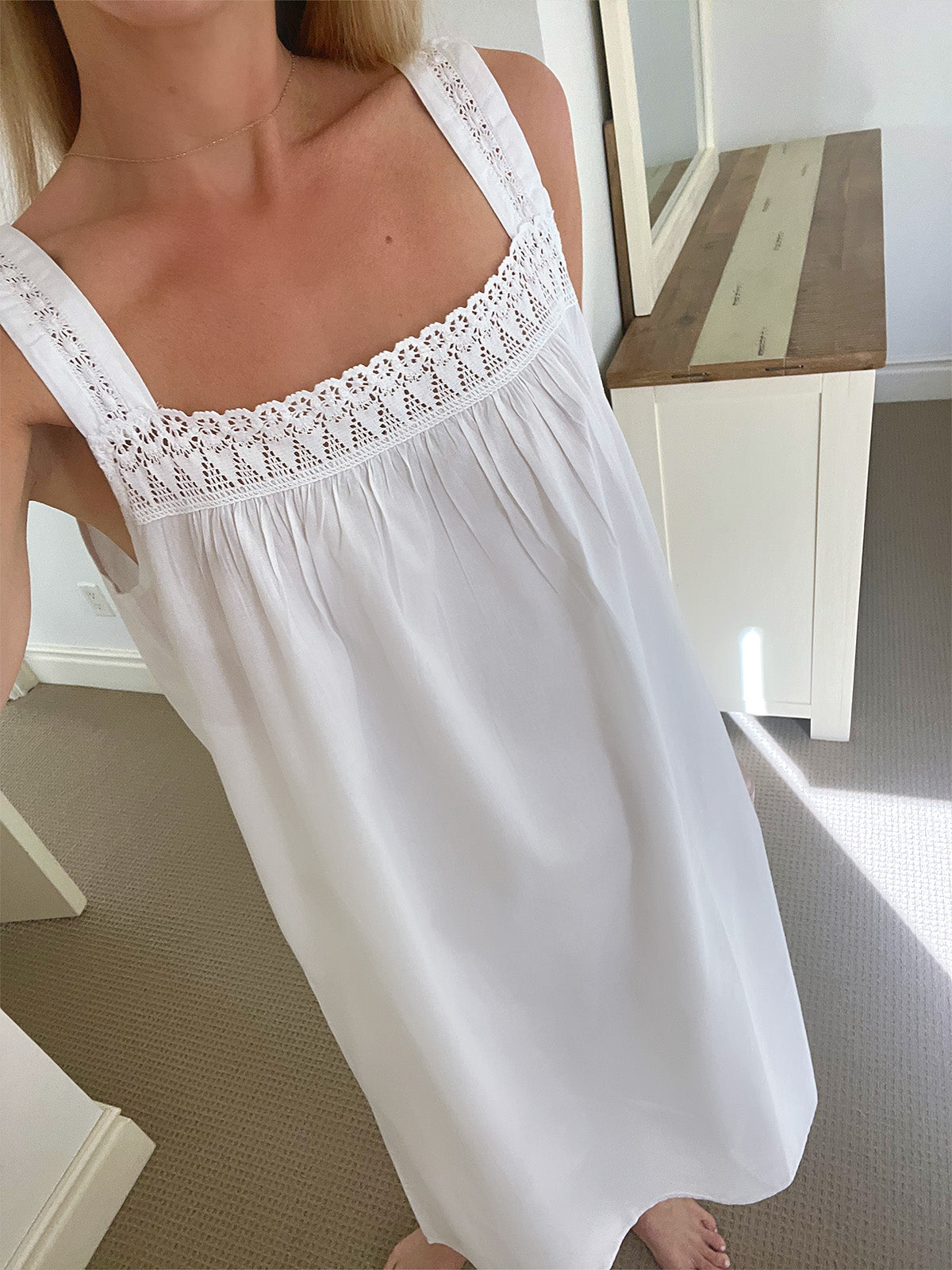 A woman wearing a white sleeveless nightgown with a square neckline and delicate lace trim, standing in a brightly lit room with a white cabinet and mirror in the background.