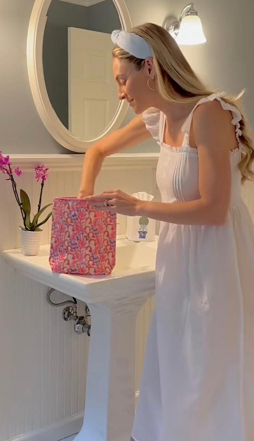 A woman in a white nightgown with ruffle sleeves standing in a bathroom, holding a pink floral bag.