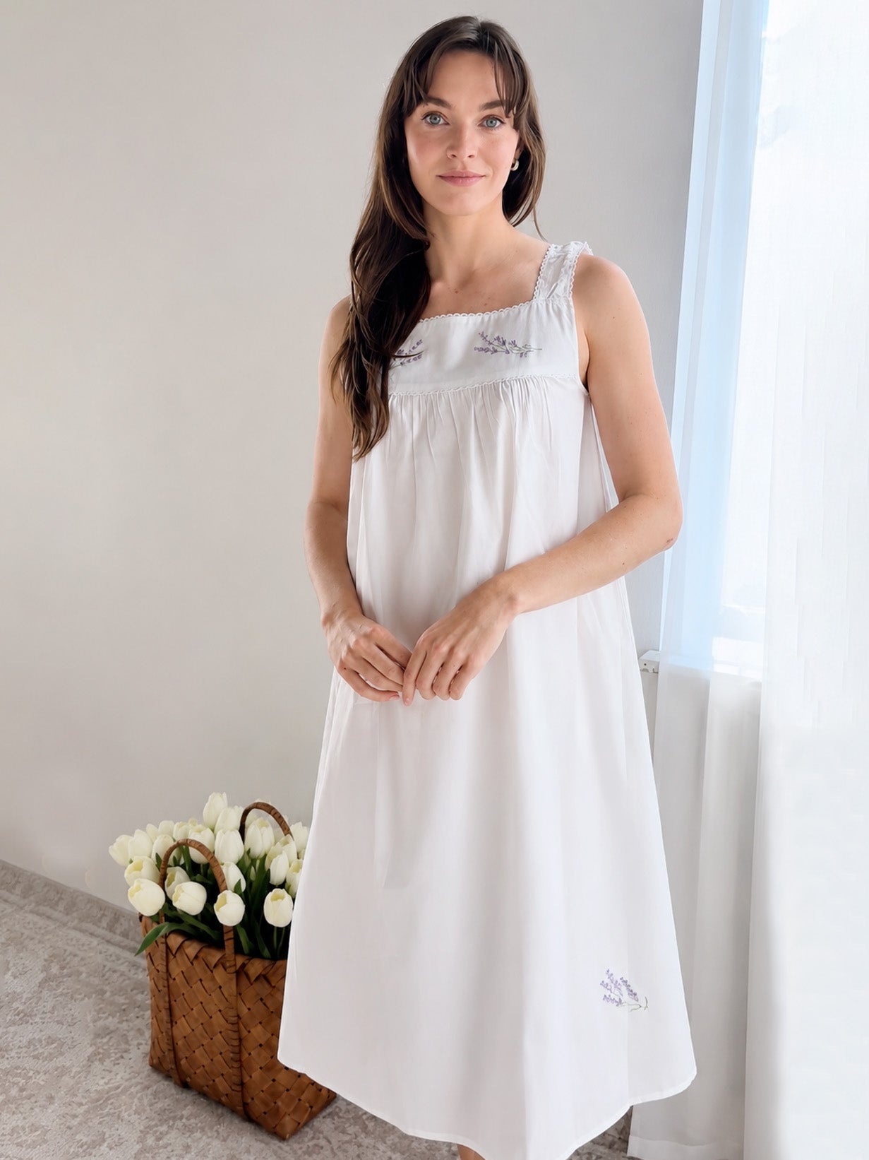 A woman wearing a white sleeveless nightgown with delicate embroidered details, standing in a brightly lit room next to a window with sheer curtains and a basket of white tulips on the floor.